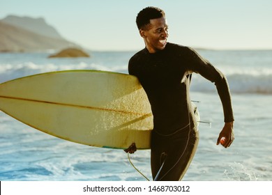 Smiling african male running out of the ocean after water surfing. Happy young man with surfboard on the beach enjoying holidays. - Powered by Shutterstock