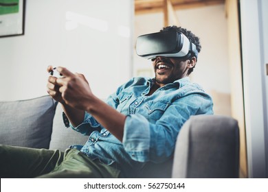 Smiling African Man Enjoying Virtual Reality Glasses While Sitting On Sofa.Happy Young Guy With Vr Headset Or 3d Spectacles And Controller Gamepad Playing Video Game At Home.Blurred