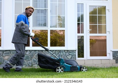 Smiling African Man In Overalls Mows The Green Grass In A Modern Garden With A Lawn Mower. A Black Man In Coveralls Pushes A Lawnmower In The Backyard. Professional Lawn Care Service.