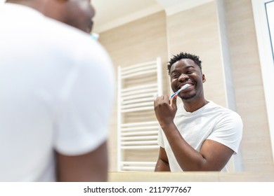 Smiling African Man With Toothbrush Cleaning Teeth And Looking Mirror In The Bathroom. Handsome Young Man Brushing His Teeth In Morning In Bathroom. Happy Guy Brushing Teeth Before Going To Sleep