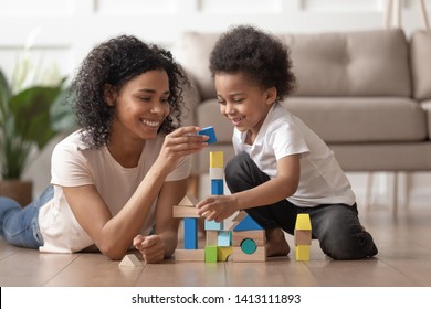 Smiling african mother baby sitter play with little kid son lay on warm floor, caring black mother nanny help teach child boy build constructor of wooden blocks at home, daycare, children development