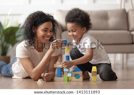 Smiling african mother baby sitter play with little kid son lay on warm floor, caring black mother nanny help teach child boy build constructor of wooden blocks at home, daycare, children development