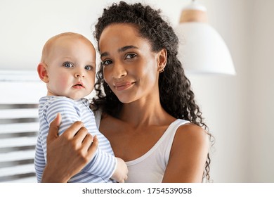 Smiling african mother holding adopted baby at home. Portrait of proud young woman rocking infant while looking at camera. Beautiful mixed race babysitter carrying adorable little child at home.