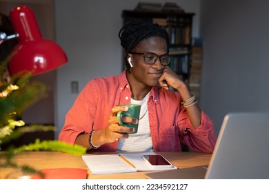 Smiling African student guy in wireless earbuds holding cup of tea coffee watching online lectures at home while studying remotely, learning online for free, looking at laptop screen. Selective focus - Powered by Shutterstock