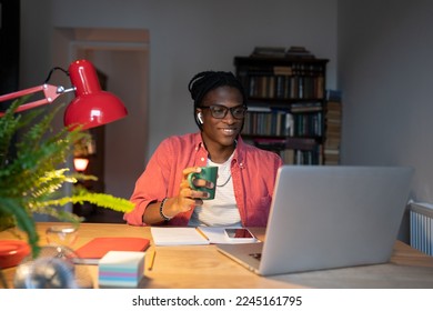 Smiling African student guy in wireless earbuds holding cup of tea coffee watching online lectures at home while studying remotely, learning online for free, looking at laptop screen. Selective focus - Powered by Shutterstock