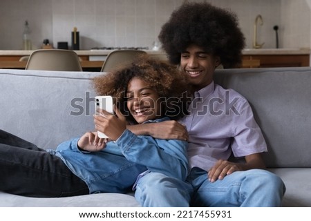 Smiling African teenager couple relaxing on sofa staring at cellphone screen feel happy enjoy new mobile application, chatting in social media, watching short-form video on hosting service. Tech, fun