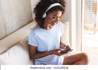 Smiling african woman in headphones using mobile phone while sitting on a couch at home: zdjęcie stockowe