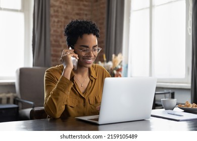 Smiling African woman sit at table looks at laptop make call by business, talk on mobile phone, provide help, do secretary job use mobile connection, make order speaks to support service, tech concept Foto Stock