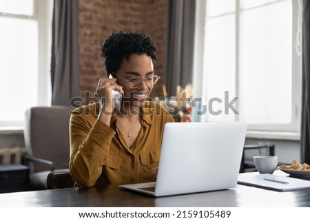 Smiling African woman sit at table looks at laptop make call by business, talk on mobile phone, provide help, do secretary job use mobile connection, make order speaks to support service, tech concept