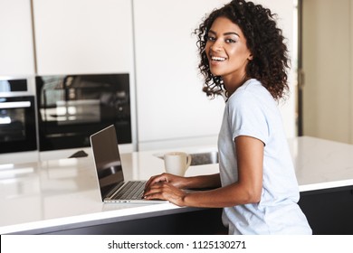 Smiling african woman using laptop computer while sitting at home with cup of coffee
