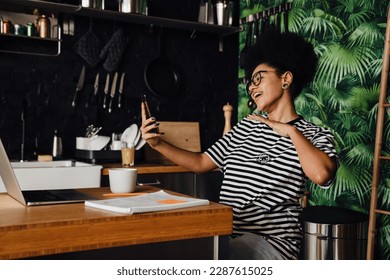 Smiling african woman wearing striped t-shirt making video call via mobile phone while working on laptop in kitchen at home - Powered by Shutterstock