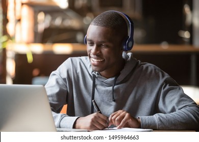 Smiling african young man student wear headphones looking at laptop screen writing notes, happy black guy e-learning in internet study online with teacher on computer watch webinar sit in cafe