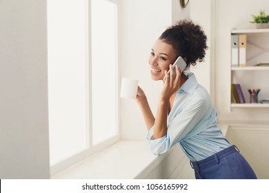 Smiling african-american business woman talking by phone near window at office, drinking coffee. Communication, technology, success concept