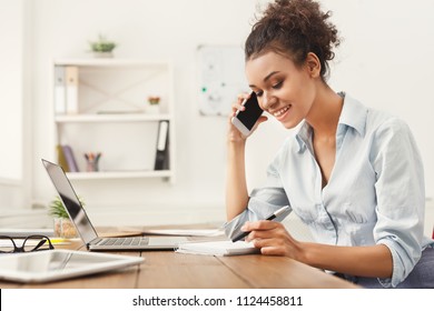 Smiling African-american Business Woman At Work Talking On Phone And Taking Notes, Sitting At Her Working Place In Office, Copy Space