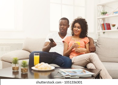 Smiling african-american couple relaxing and watching TV at home, changing channels, having rest after hard week, copy space