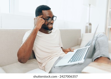 Smiling AfricanAmerican Freelancer Working on Laptop in Modern Home Office This image features a young AfricanAmerican man sitting on a comfortable sofa in his stylish living room He is happily typing