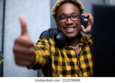 Smiling African-American gamer with headphones liked the game and he gives a thumbs-up gesture. A black guy in a plaid shirt and with an African hairstyle enjoys the game and shares emotions on camera