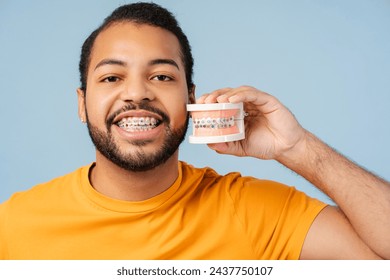 Smiling, african-american man with braces, holding dental mold with braces, smiling, isolated on blue background. Emphasizes orthodontic therapy concept  - Powered by Shutterstock