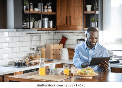 Smiling African-American man reading on a tablet while having breakfast at home. He is enjoying a healthy meal and coffee in a bright, modern kitchen. - Powered by Shutterstock