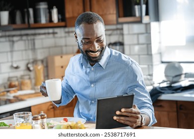 Smiling African-American man reading on a tablet while having breakfast at home. He is enjoying a healthy meal and coffee in a bright, modern kitchen. - Powered by Shutterstock