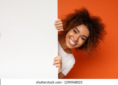 Smiling African-american Woman Looking Out Of White Blank Sign With Copy Space On Orange Background