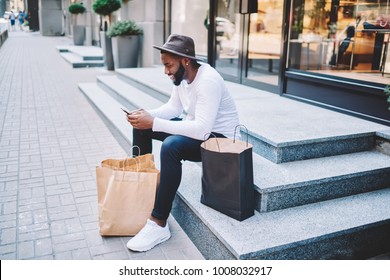 Smiling Afro American Male Customer In Trendy Hat Sitting On Stairs Of Store Waiting For Delivery Service Sending Email On Mobile, Dark-skinned Guy Satisfied With Online Shopping Resting With Bags