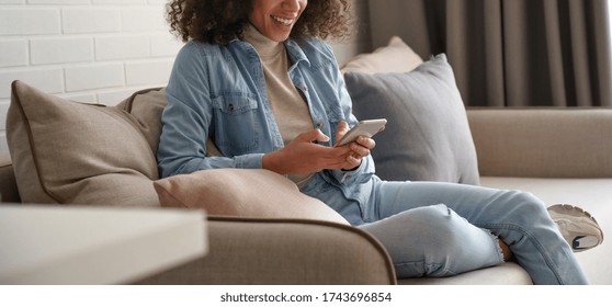 Smiling Afro American Millennial Woman User Holding Smart Phone Watching Video, Playing Game, Shopping, Chatting In Mobile Technology Application Sitting On Sofa At Home. Close Up View, Banner Design.