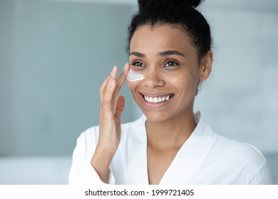 Smiling Afro American woman in bathrobe spreading moisturizing collagen cream on face, applying mask on under eye skin for reducing wrinkles, dryness, removing bags. Beauty care, skincare concept - Powered by Shutterstock