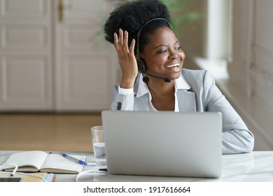 Smiling Afro American Woman Call Center Agent Wearing Headset With Microphone Talking With Customer, Looking At Window. Happy Black Girl Dispatcher In Earphones Laughing, Working On Laptop In Office