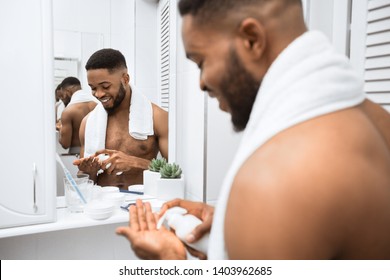 Smiling afro man with naked torso squeezing cream on his hand, reflecting in mirror at bathroom. Man's skin care concept