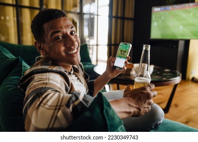 Smiling afro man watching football match and making bets at bookmaker's website while sitting on sofa in front of TV screen - Shutterstock ID 2230349743