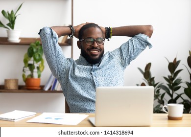 Smiling Afro-American businessman holding hands behind head sitting at office desk behind laptop. Happy black employee feeling no stress, relaxing, watching funny video after successful working - Shutterstock ID 1414318694
