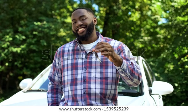 Smiling afro-american man showing car keys\
leaning new automobile, rent\
service