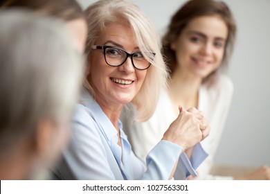 Smiling aged businesswoman in glasses looking at colleague at team meeting  happy attentive female team leader listening to new project idea  coach mentor teacher excited by interesting discussion
