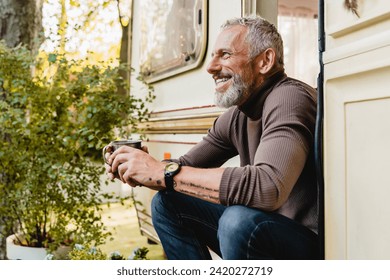 Smiling aged handsome man with beard and tattoo sitting near his trailer holding a mug of coffee, having breakfast while driving traveling alone by camper van motor home - Powered by Shutterstock