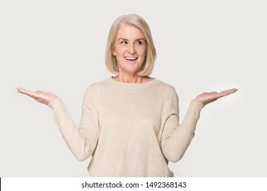 Smiling Aged Senior Lady Isolated On Grey Studio Background Hold Hands Aside Compare Two Options, Happy Positive Mature Woman Make Choice Decide Evaluate Offer Or Deal, Consider Pros And Cons