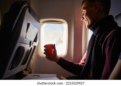 Smiling Airline Passenger With A Paper Cup Of Coffee Looking Ahead In Airplane Window. Man Drinks Tea Onboard. Male Holds Beverage Or Water During Flight.