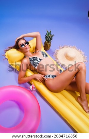  smiling amazing beautiful caucasian female who wears in bikini lyes on inflatable yellow matress while holds cocktail on purple background . Summer concept. Travel concept. Relax concept