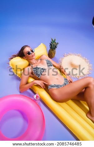  smiling amazing beautiful caucasian girl who wears in bikini lyes on inflatable yellow matress while holds cocktail on purple background . Summer concept. Travel concept. Relax concept