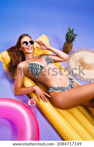  smiling amazing beautiful caucasian woman who wears in bikini lyes on inflatable yellow matress while posing on purple background . Summer concept. Travel concept. Relax concept