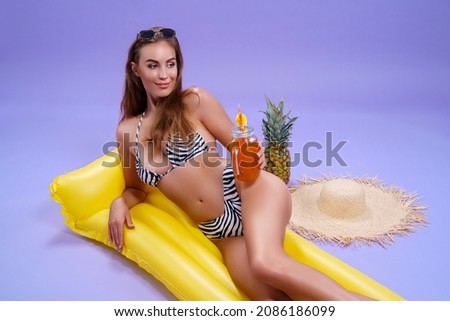  smiling amazing beautiful caucasian woman who wears in bikini lyes on inflatable yellow matress while holds cocktail on purple background . Summer concept. Travel concept. Relax concept