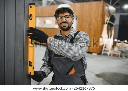 Smiling arab builder checking vertical alignment of modular house wall with spirit level in prefabricated house workshop