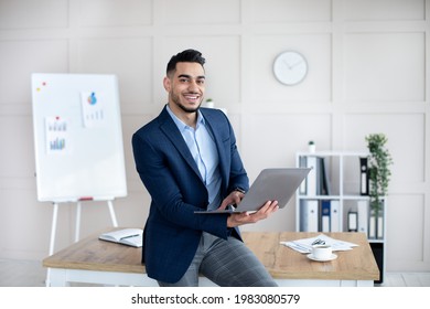 Smiling Arab Businessman In Formal Suit Holding Laptop Computer At Contemporary Office. Young Male CEO Conducting Online Company Meeting Or Conference With Coworkers, Indoors