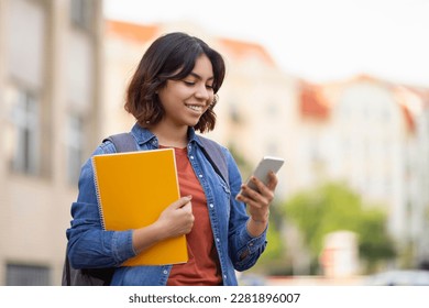 Smiling Arab College Student Using Smartphone And Holding Workbooks While Resting Outdoors After Classes, Cheerful Young Middle Eastern Woman Texting On Mobile Phone Or Browsing New App, Copy Space