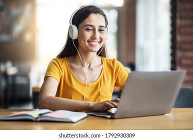 Smiling arab girl with headset studying online, using laptop at cafe, taking notes, copy space