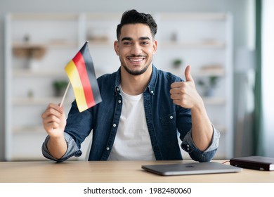 Smiling arab guy with flag of Germany showing thumb up, sitting at desk with laptop, middle-eastern young man student learning German for job, education or emmigration to Germany, copy space - Shutterstock ID 1982480060