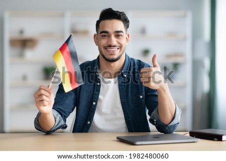 Smiling arab guy with flag of Germany showing thumb up, sitting at desk with laptop, middle-eastern young man student learning German for job, education or emmigration to Germany, copy space
