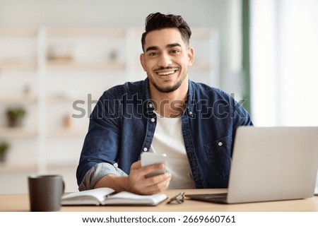 Smiling arab guy using smartphone while sitting at workdesk