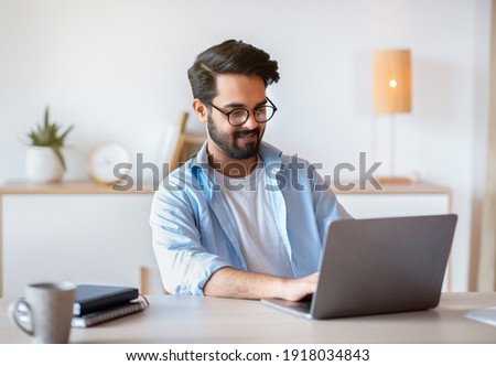 Smiling Arab Man Self-Entrepreneur Working With Laptop At Home Office, Sitting At Desk And Using Computer For Online Project, Eastern Male Freelancer Enjoying Distant Job Opportunities, Copy Space