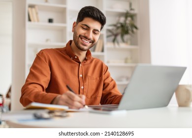 Smiling Arab Man Using Laptop Working With Documents At Home Office, Handsome Eastern Male Entrepreneur Sitting At Desk With Computer, Checking Financial Reports And Taking Notes, Free Copy Space